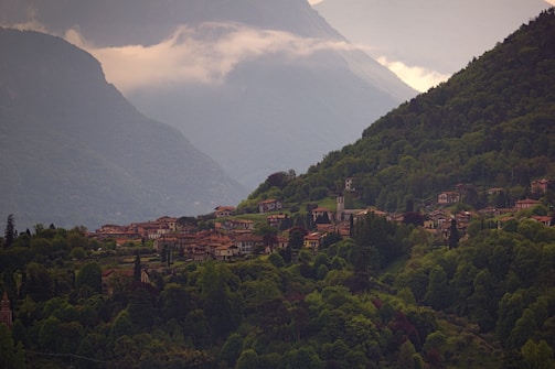 A lush green valley enveloped by tall mountains is dotted with quaint houses. The village is nestled amidst dense foliage, with a few towers and buildings standing out. Mist hovers over the mountaintops, adding a serene and tranquil atmosphere to the landscape.