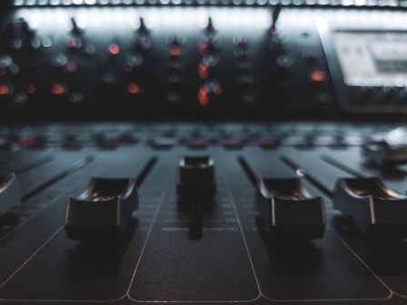 A close-up view of a mixing console with several faders and knobs, set in a dimly lit environment. The background displays a soft blur of colorful lights and dials, suggesting an active audio studio setup.