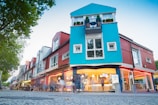 A vibrant urban scene featuring a modern building with a unique architectural design. The structure is painted in bright colors, predominantly blue and red, with large windows and greenery on the balconies. The ground floor has a cafe or restaurant with outdoor seating and people gathered around, creating a lively atmosphere.