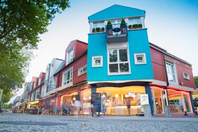 A vibrant urban scene featuring a modern building with a unique architectural design. The structure is painted in bright colors, predominantly blue and red, with large windows and greenery on the balconies. The ground floor has a cafe or restaurant with outdoor seating and people gathered around, creating a lively atmosphere.