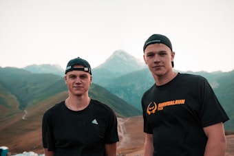 Two young men wearing black sports attire and caps stand in front of a scenic mountainous landscape. The mountains in the background have a mix of green slopes and rocky peaks, with a clear sky overhead. Both individuals have a relaxed expression.