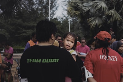 A group of people gathered outdoors, featuring a man holding a young child, who appears expressive. The group includes people wearing various colorful shirts, such as pink and orange, with some in sports attire like hockey shirts. The background consists of lush greenery and trees.