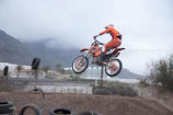 Rider performing a jump on a pitbike with orange accents in a forest setting