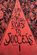 A vibrant street art mural with a central red pathway containing black text that reads, 'Life is a straight road to success.' Surrounding the pathway are intricate black and white drawings of smiling faces and abstract shapes, creating a lively and optimistic atmosphere.