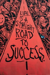 A vibrant street art mural with a central red pathway containing black text that reads, 'Life is a straight road to success.' Surrounding the pathway are intricate black and white drawings of smiling faces and abstract shapes, creating a lively and optimistic atmosphere.