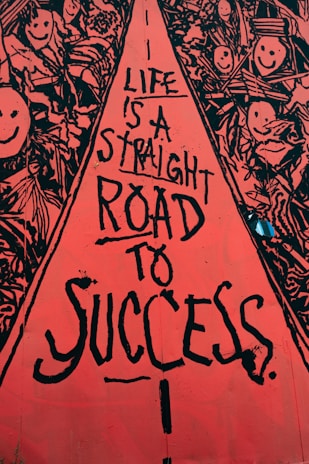 A vibrant street art mural with a central red pathway containing black text that reads, 'Life is a straight road to success.' Surrounding the pathway are intricate black and white drawings of smiling faces and abstract shapes, creating a lively and optimistic atmosphere.