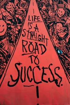 A vibrant street art mural with a central red pathway containing black text that reads, 'Life is a straight road to success.' Surrounding the pathway are intricate black and white drawings of smiling faces and abstract shapes, creating a lively and optimistic atmosphere.