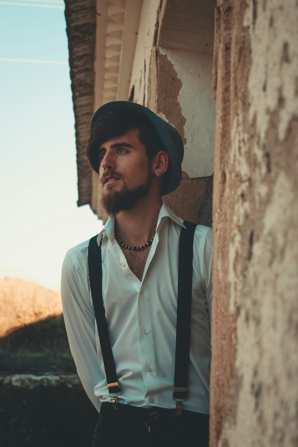 man wearing white button-up dress shirt and suspenders leaning on brown and white concrete wall