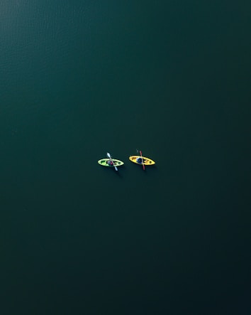 Two kayaks, one green and one yellow, are floating on a vast expanse of dark green water. The kayaks are occupied by individuals who appear to be paddling calmly side by side.
