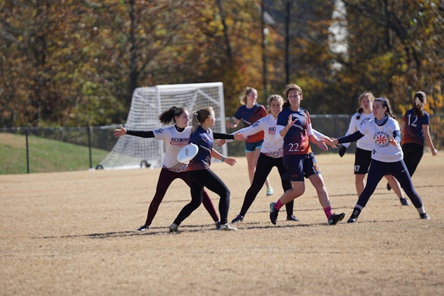 A group of people is playing ultimate frisbee on a grassy field. They are in motion, with one player holding the frisbee while others attempt to block or receive. Trees and a soccer goal can be seen in the background.