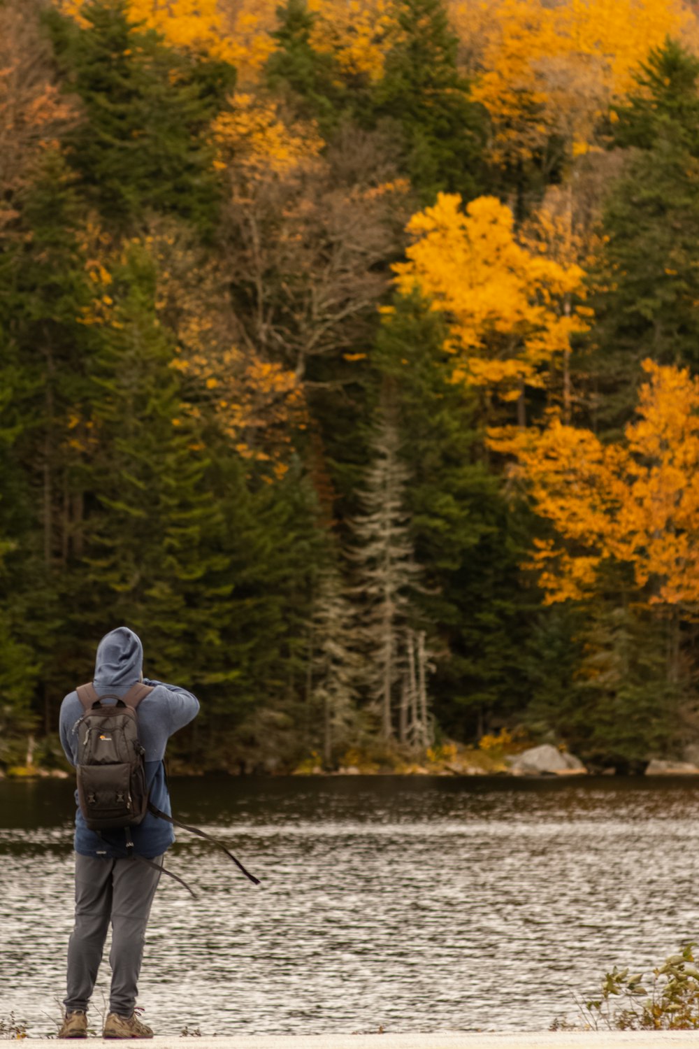 Homme en sweat à capuche bleu et sac à dos noir près d’un plan d’eau pendant la journée