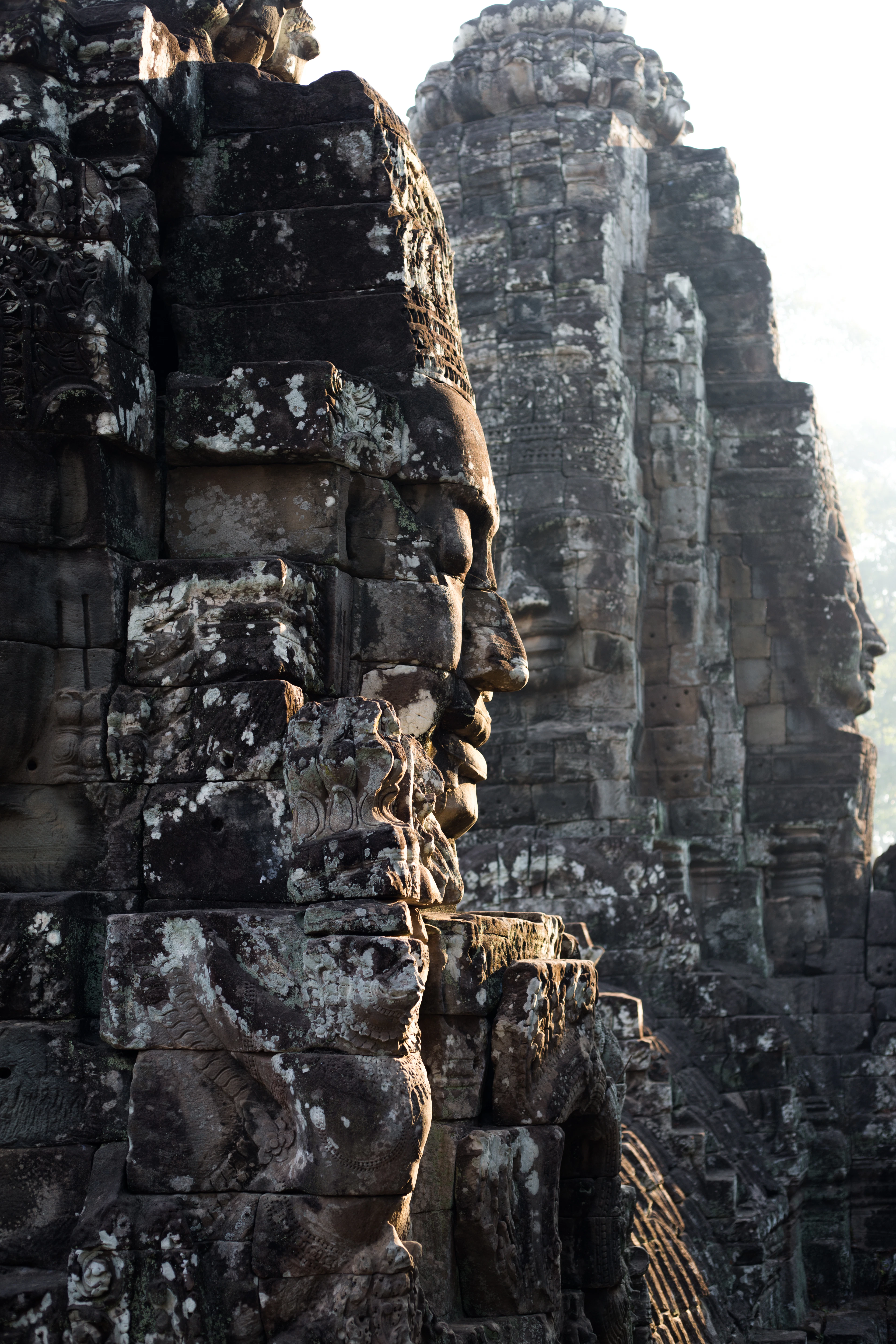 Colossal carved stone faces on the lichen-covered towers of the Bayon temple at Angkor Thom, seen in profile against morning haze