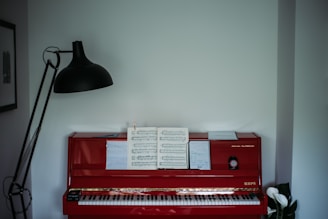 A red upright piano with sheet music and a notebook placed on its music stand. A tall black floor lamp stands on one side, casting light towards the piano. On the other side, there is a small plant with white flowers. The room has minimal decor with a framed picture visible on the wall.