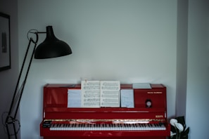 A red upright piano with sheet music and a notebook placed on its music stand. A tall black floor lamp stands on one side, casting light towards the piano. On the other side, there is a small plant with white flowers. The room has minimal decor with a framed picture visible on the wall.