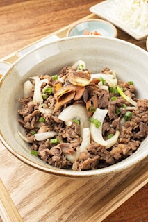 A bowl filled with a savory dish featuring sliced beef, garnished with onions, green onions, and crispy garlic chips. The bowl is placed on a wooden surface, and there are small side dishes in the background.
