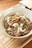 A bowl filled with a savory dish featuring sliced beef, garnished with onions, green onions, and crispy garlic chips. The bowl is placed on a wooden surface, and there are small side dishes in the background.