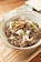 A bowl filled with a savory dish featuring sliced beef, garnished with onions, green onions, and crispy garlic chips. The bowl is placed on a wooden surface, and there are small side dishes in the background.