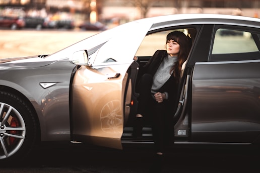 A person is seated inside a sleek, modern car with the door open, partially stepping out. The car is parked in a spacious, sunlit area. The individual is dressed in a casual but stylish manner, wearing a dark coat over a light-colored sweater. The car’s exterior is shiny and appears to be a luxury model.
