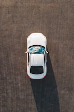 A top-down view of a white car parked on a large expanse of asphalt, with a reflection of clouds visible on its sunroof.