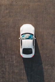 A top-down view of a white car parked on a large expanse of asphalt, with a reflection of clouds visible on its sunroof.