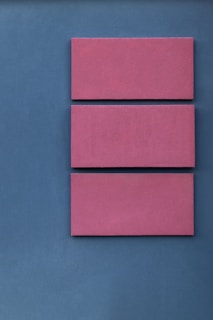 Three pink rectangular panels are arranged vertically and evenly spaced on a textured blue background. The simplicity and geometric alignment create a minimalist aesthetic.