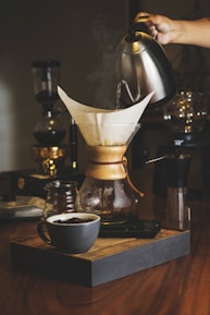 A hand is pouring hot water from a kettle into a Chemex coffee maker with a paper filter, creating a warm and cozy atmosphere. The setup includes a cup of coffee beans on a wooden tray, set on a wooden table with additional coffee-making equipment in the background.