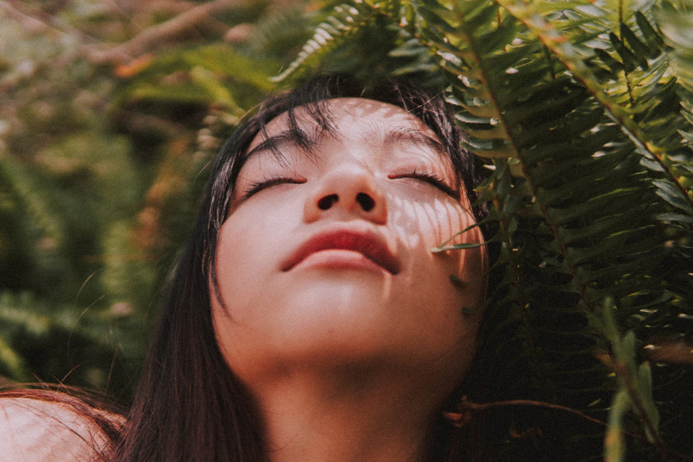 A young woman with her eyes closed, face turned up to the sun among green fern leaves