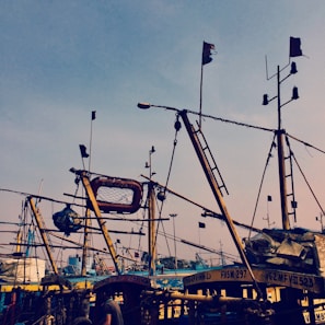 A cluster of fishing boats with wooden masts and various equipment are closely docked together. Flags flutter atop the masts, and the sky serves as a backdrop. A life preserver and several ropes are draped across the boats, giving a sense of preparedness for sea travel.