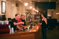 A cozy coffee shop with warm lighting features four people interacting. Three are standing behind a wooden counter, busy with drinks, while one person stands on the opposite side, possibly a customer. The ambiance suggests a casual and relaxed environment with potted plants, shelves, and hanging light bulbs adding to the decor.
