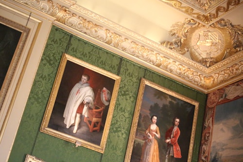 A richly decorated interior wall featuring ornate gold moldings and an elaborate crest. The wall is adorned with several framed paintings. The paintings depict historical figures in period attire. The background features a luxurious green patterned wallpaper.