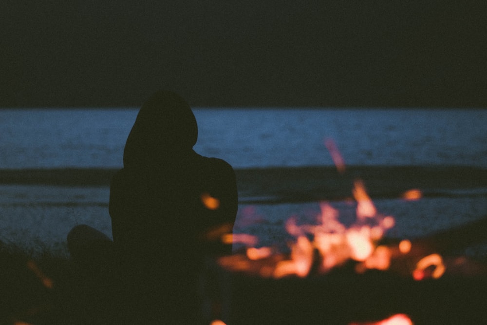 silhouette of woman sitting on seashore near bonfire
