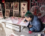 A person with green hair and a blue jacket is focused on creating intricate artwork at a low table, surrounded by various detailed and colorful prints that lean against a wall. The artwork features complex patterns with red, black, and white colors prominent.