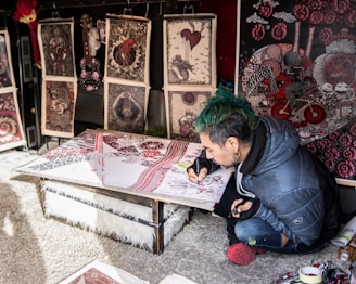 A person with green hair and a blue jacket is focused on creating intricate artwork at a low table, surrounded by various detailed and colorful prints that lean against a wall. The artwork features complex patterns with red, black, and white colors prominent.
