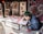 A person with green hair and a blue jacket is focused on creating intricate artwork at a low table, surrounded by various detailed and colorful prints that lean against a wall. The artwork features complex patterns with red, black, and white colors prominent.