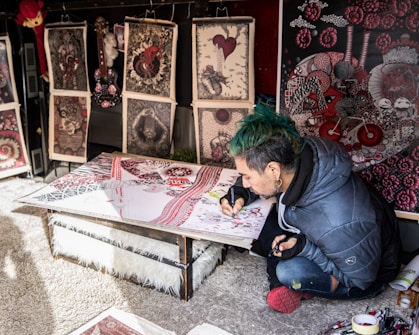 A person with green hair and a blue jacket is focused on creating intricate artwork at a low table, surrounded by various detailed and colorful prints that lean against a wall. The artwork features complex patterns with red, black, and white colors prominent.