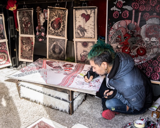 A person with green hair and a blue jacket is focused on creating intricate artwork at a low table, surrounded by various detailed and colorful prints that lean against a wall. The artwork features complex patterns with red, black, and white colors prominent.