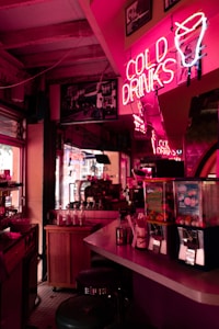 A dimly lit bar or diner area with a vibrant neon sign reading 'Cold Drinks', along with a neon ice cream cone illustration. The counter is equipped with several blenders, syrup dispensers, and gumball machines. Bar stools line the counter, and a cozy, vintage atmosphere is created by the warm, pink lighting and retro decor.