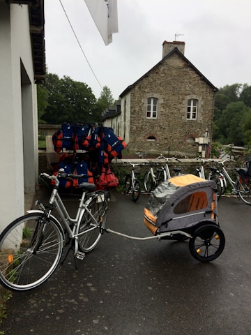A bicycle with a child trailer attached is parked near a building with brick and stone walls. Several other bicycles and a rack filled with life jackets are visible nearby. The area appears to be set in a rustic, possibly rural, setting with trees in the background.