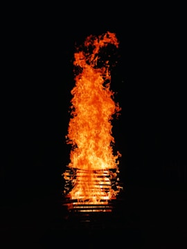 A large, bright orange flame rises dramatically from a stack of wooden pallets against a dark background. The fire appears intense and is surrounded by swirling patterns of smoke.