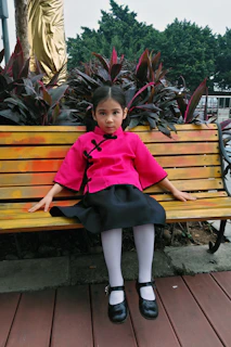 A little girl sitting gracefully on a vintage wooden bench wearing a sharara set with subtle gold accents, surrounded by festive lanterns.