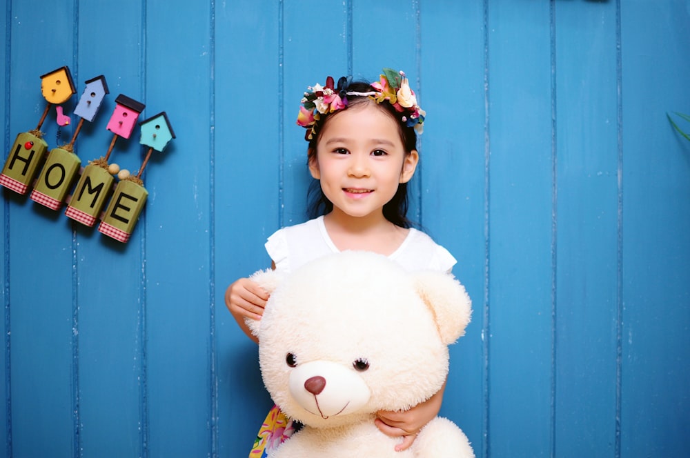 girl with white teddy bear stands with back on blue wooden wall