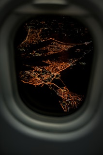 A nighttime aerial view of a cityscape with networks of illuminated streets and clusters of lights visible through an airplane window frame. The winding streets form intricate patterns against the dark backdrop of the night.