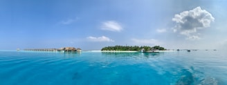A tranquil ocean scene with crystal clear blue water extending to an overwater bungalow resort on the left. A lush, green island with palm trees and white sandy beach occupies the center, with a few boats anchored nearby. The sky is clear with a few fluffy clouds.