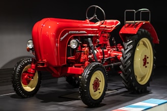 A vintage red tractor with a classic design and large black tires displayed indoors, featuring smooth curves and a prominent steering wheel. The tractor is illuminated against a dark background, highlighting its polished exterior and mechanical details, including the exposed engine components and vertical exhaust.