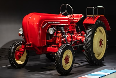 A vintage red tractor with a classic design and large black tires displayed indoors, featuring smooth curves and a prominent steering wheel. The tractor is illuminated against a dark background, highlighting its polished exterior and mechanical details, including the exposed engine components and vertical exhaust.