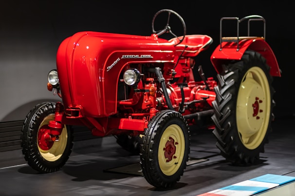 A vintage red tractor with a classic design and large black tires displayed indoors, featuring smooth curves and a prominent steering wheel. The tractor is illuminated against a dark background, highlighting its polished exterior and mechanical details, including the exposed engine components and vertical exhaust.