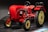 A vintage red tractor with a classic design and large black tires displayed indoors, featuring smooth curves and a prominent steering wheel. The tractor is illuminated against a dark background, highlighting its polished exterior and mechanical details, including the exposed engine components and vertical exhaust.