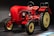 A vintage red tractor with a classic design and large black tires displayed indoors, featuring smooth curves and a prominent steering wheel. The tractor is illuminated against a dark background, highlighting its polished exterior and mechanical details, including the exposed engine components and vertical exhaust.