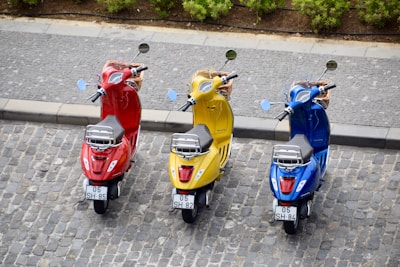 Three scooters are parked on a cobblestone street, each in a different color: red, yellow, and blue. The background shows a sidewalk, and there are green shrubs beyond the road.