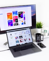 A modern workspace featuring a MacBook Pro with open design software and a large monitor displaying colorful design reports. A black mug, a potted plant, and a smartphone are also visible on the white desk. A Starbucks cup is placed near the monitor.